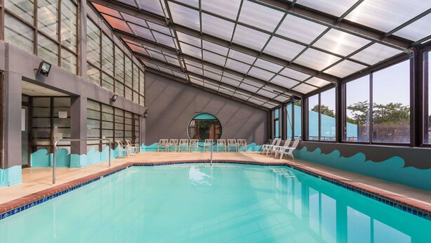 An indoor swimming pool with a glass ceiling, clear poolside chairs, and a view of the outdoors. - Howard Johnson by Wyndham Williamsburg (Williamsburg)