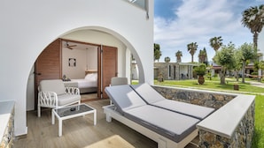 Un espacio lounge moderno al aire libre con sofá, sillas y una tumbona, con vistas a un jardín cuidado con palmeras.
