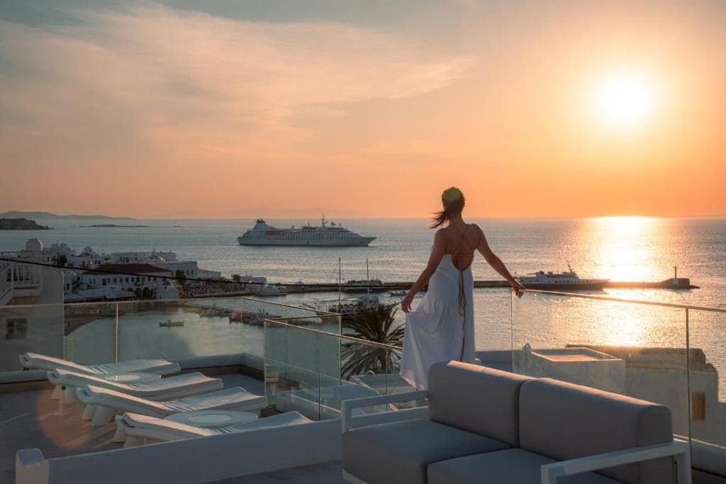 Eine Person steht auf einem Balkon und blickt auf einen Sonnenuntergang, im Hintergrund ist ein Kreuzfahrtschiff zu sehen.