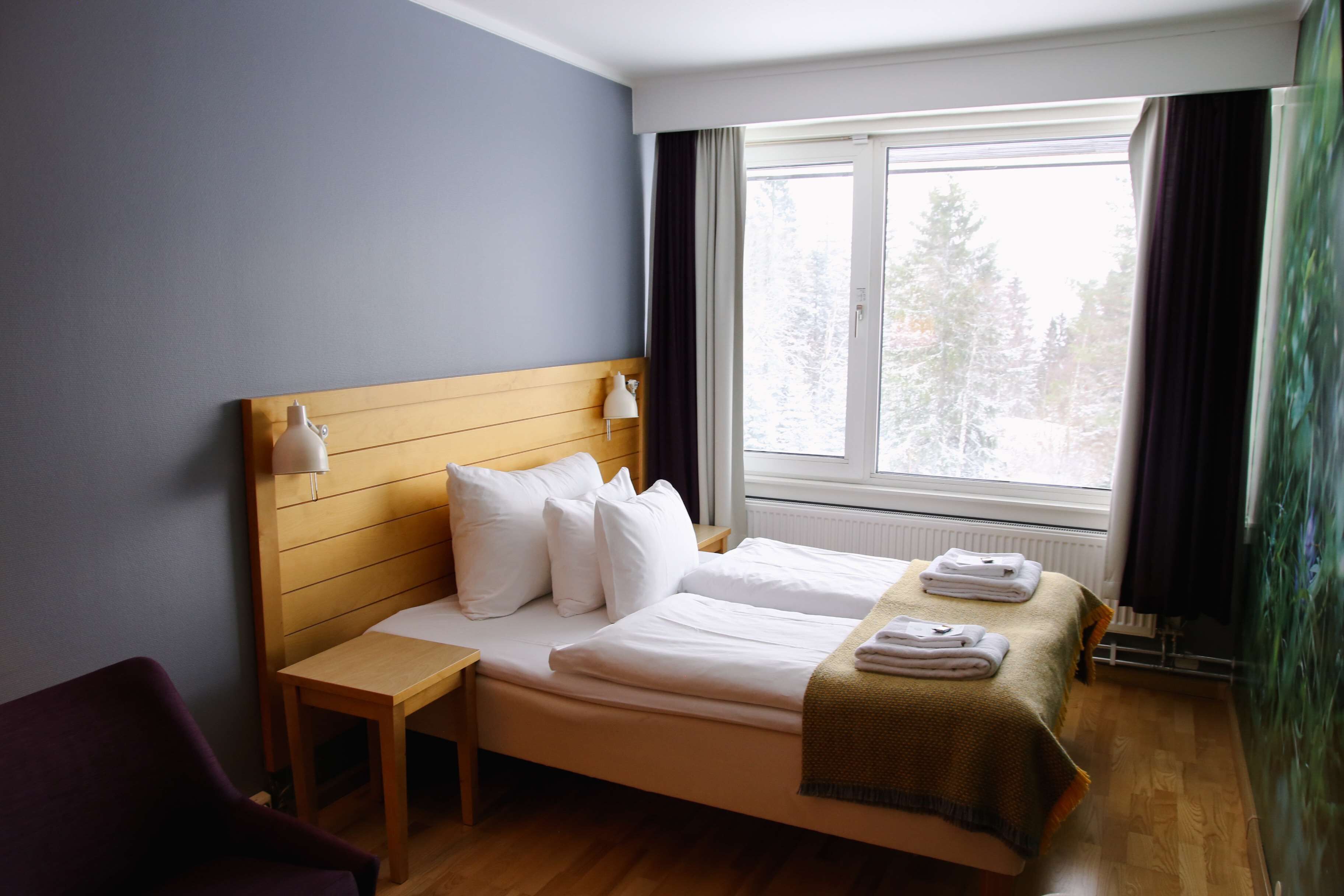 A hotel room with a wooden headboard, a single bed with white linens, a nightstand, a chair, and a window with curtains.