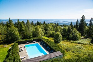 A rectangular swimming pool surrounded by a wooden deck, with a view of a forested area and a distant cityscape. - Voksenasen Hotell, BW Signature Collection (Oslo)