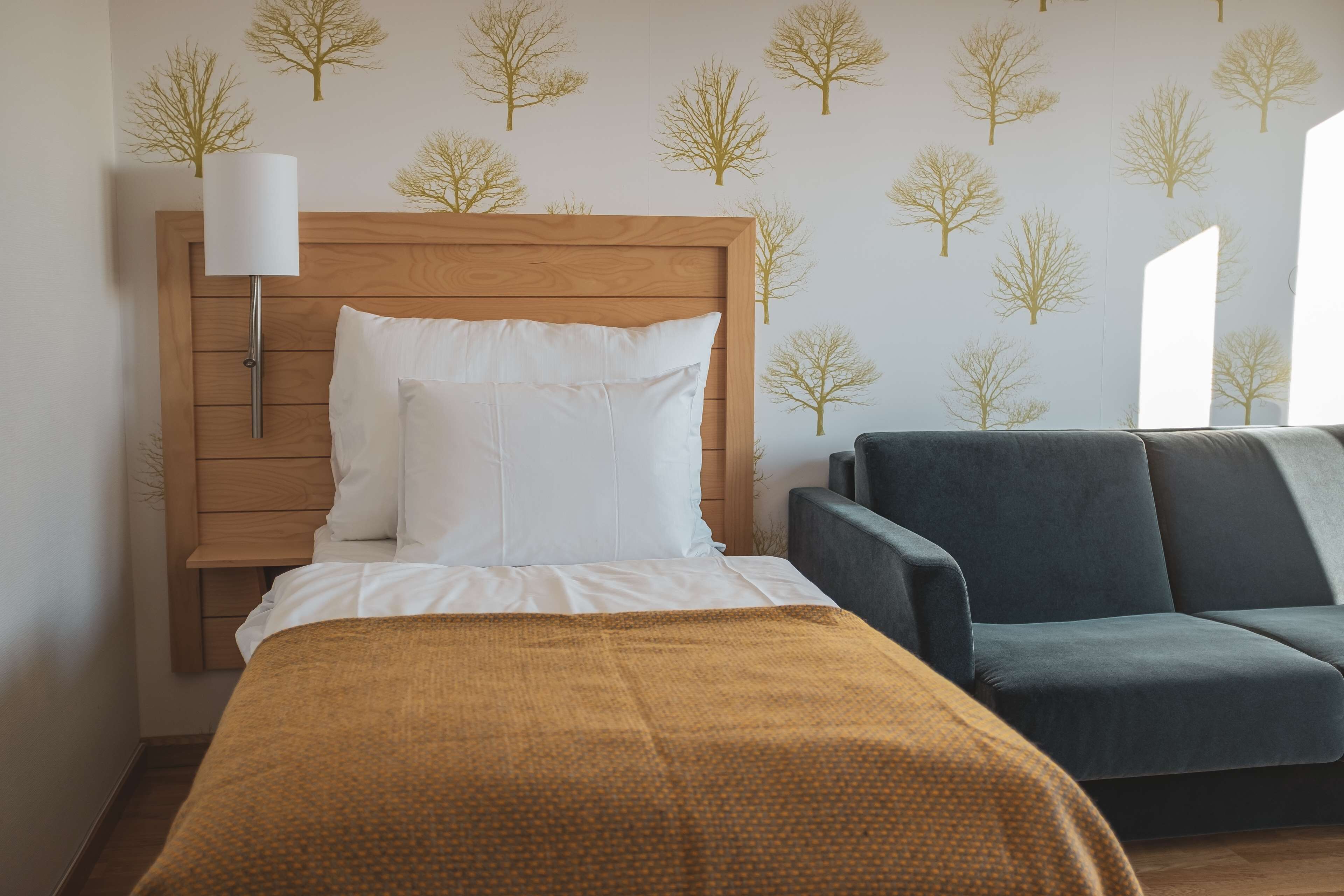 Una habitación de hotel con cabecera de madera, cama con ropa blanca, manta color mostaza y sofá azul oscuro.
