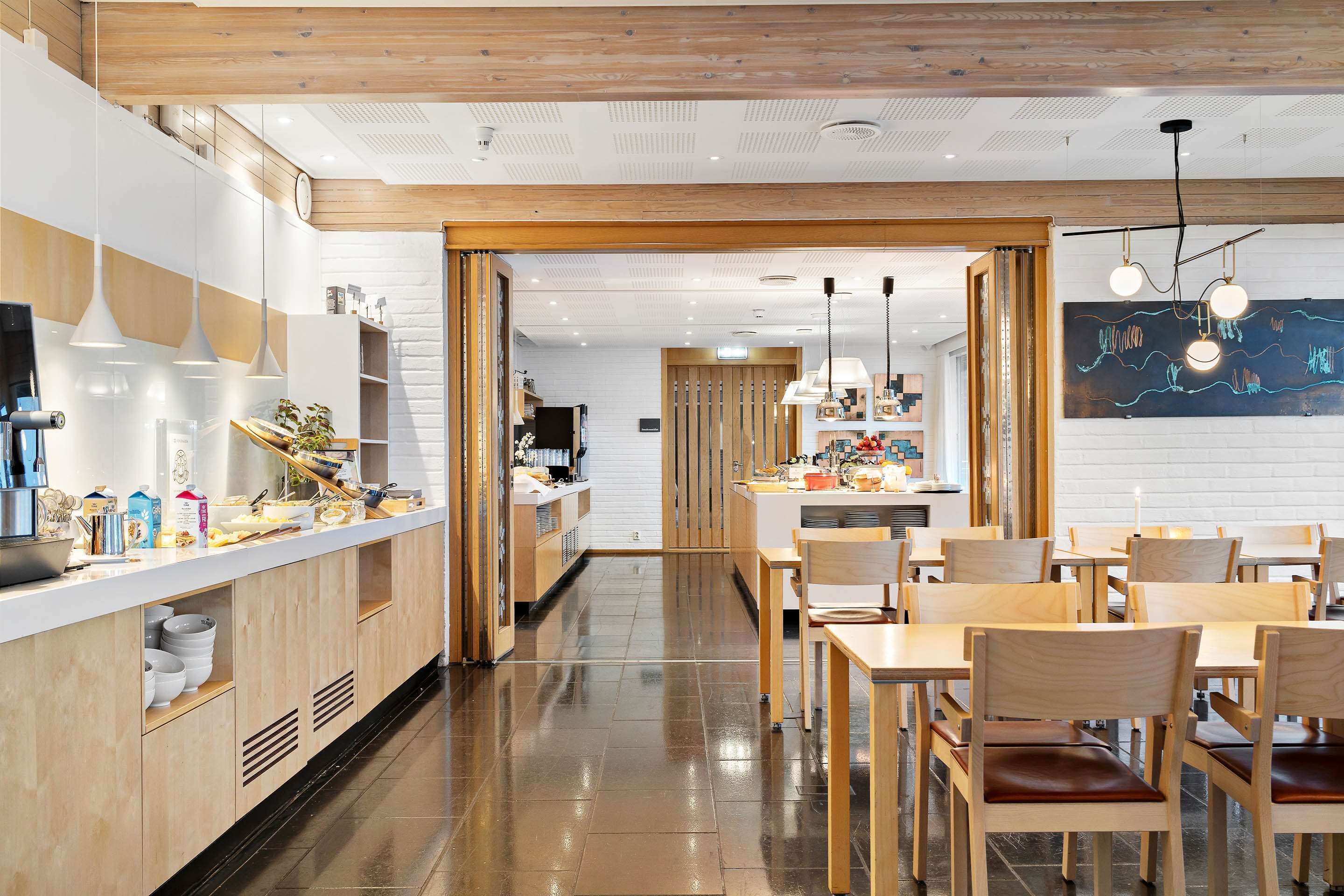 Un comedor moderno con mesas y sillas de madera, una cocina al fondo e iluminación colgante.