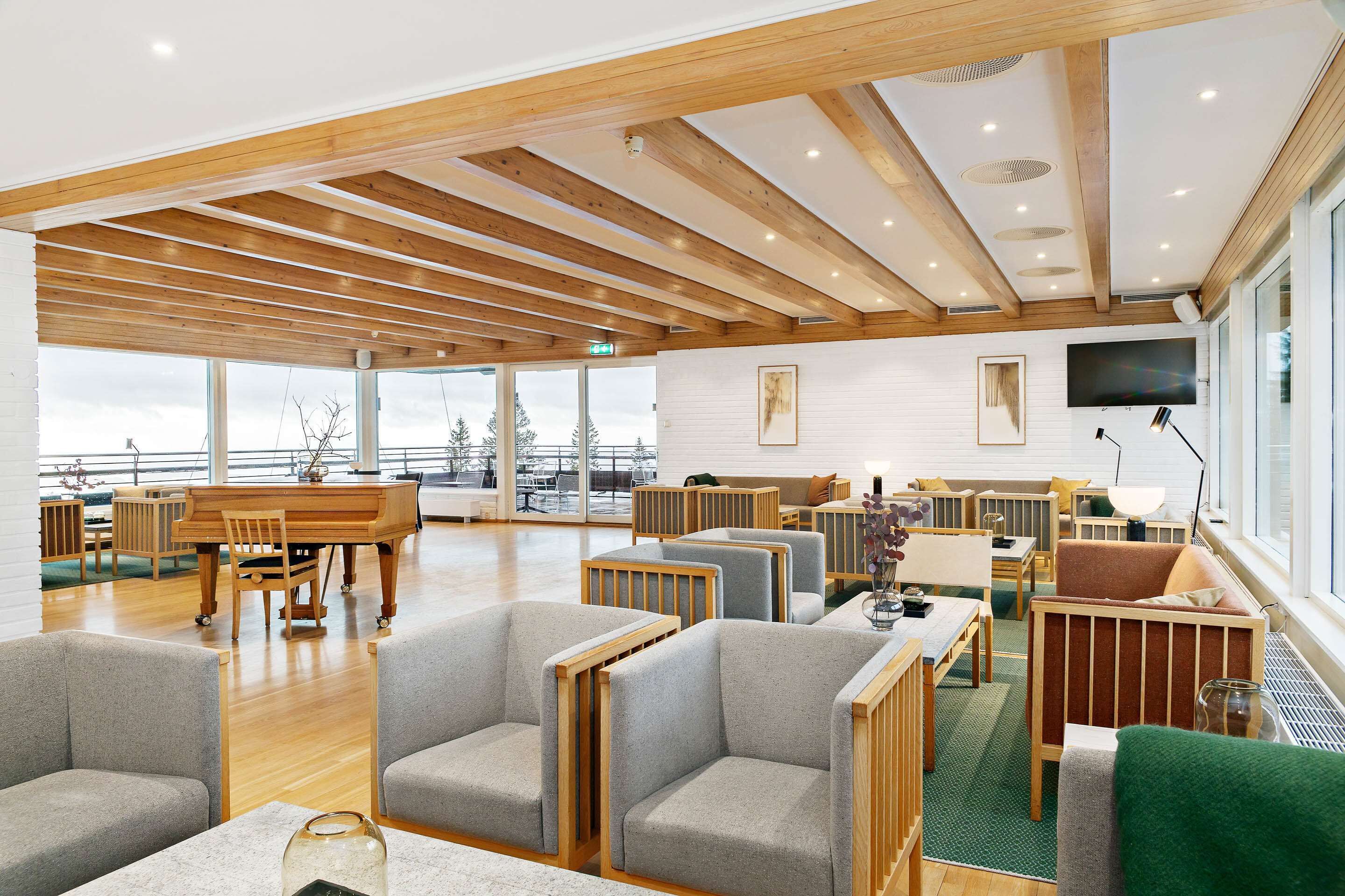 A spacious lounge area with wooden beams, a piano, comfortable seating, and large windows offering views.