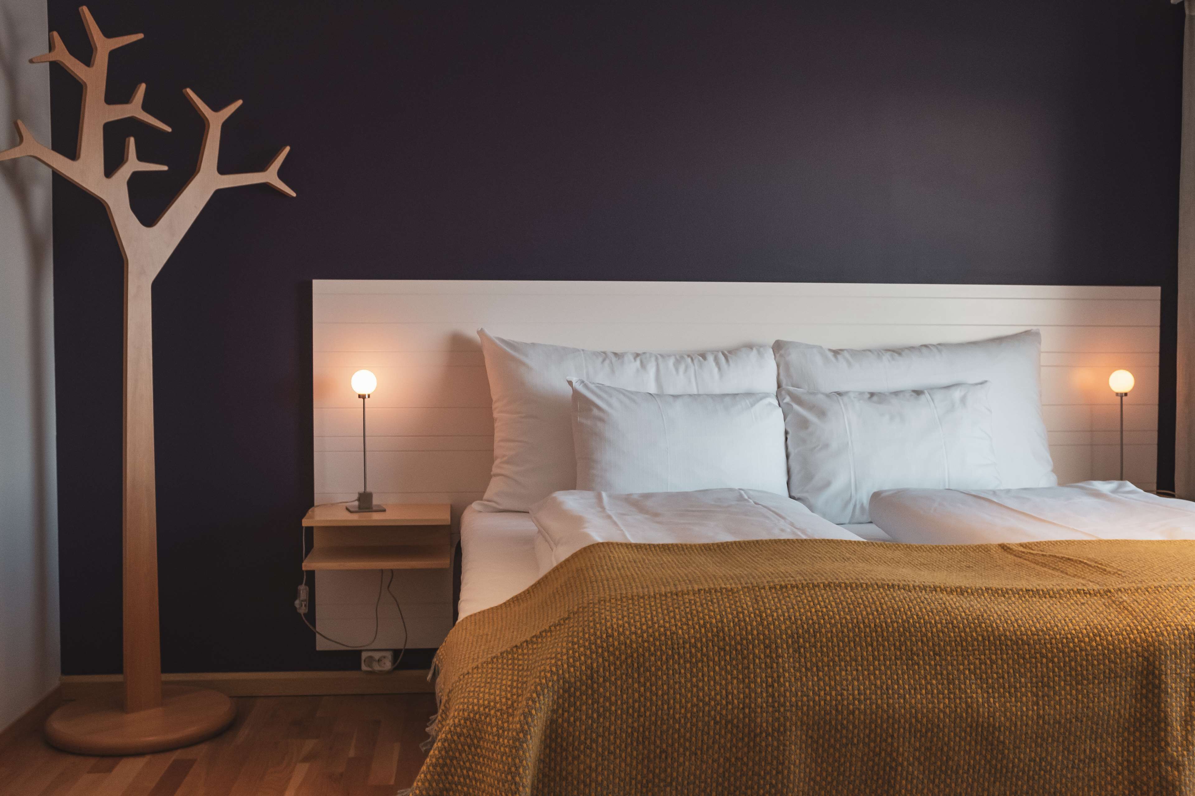 Un dormitorio con una cama blanca, dos lámparas de noche, un adorno con forma de árbol de madera y una pared oscura.
