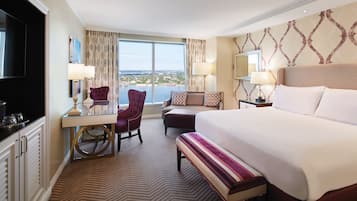 A hotel room with a large bed, a desk with a chair, a television, and a view of the city.