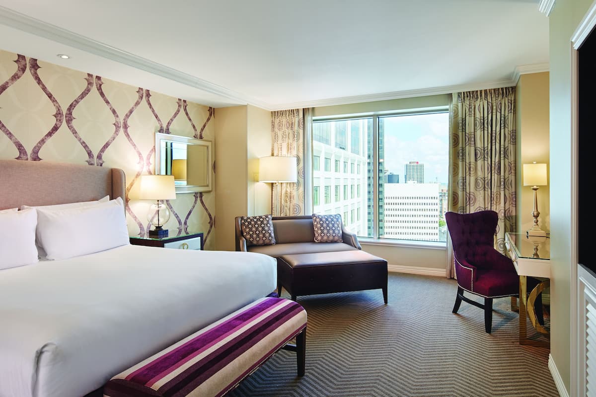 A hotel room with a large bed, a sofa, a chair, and a desk with a lamp. There is a window with curtains and a view of buildings outside.