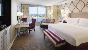 A hotel room with a large bed, a desk with a chair, a television, and a view of the city through large windows.