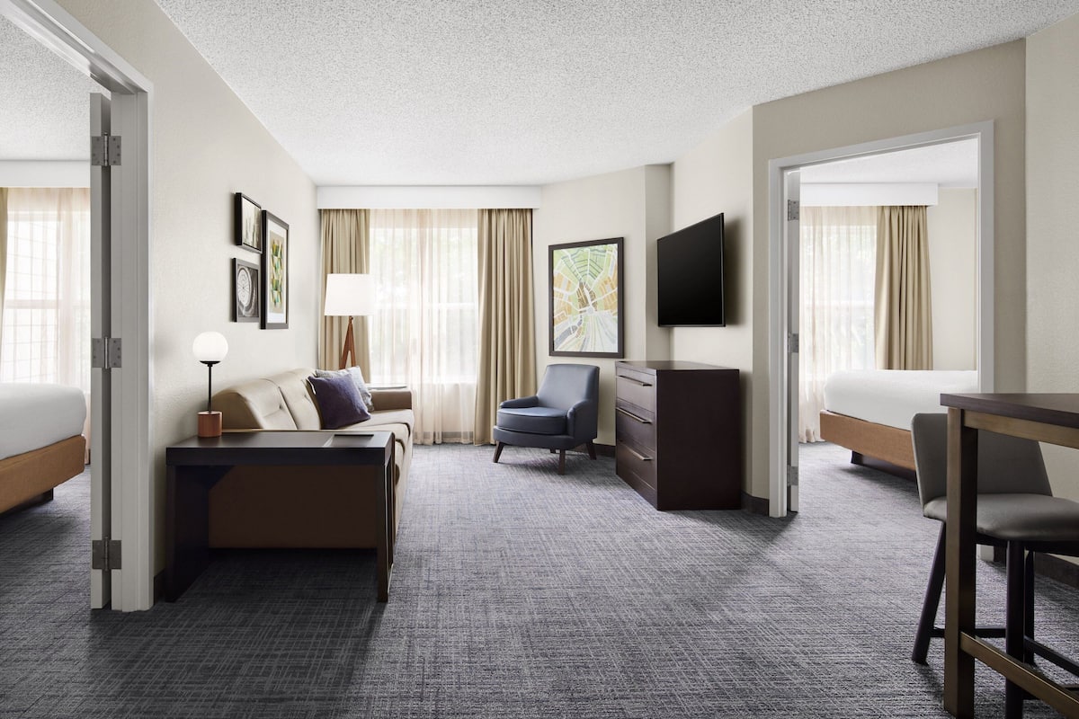 A hotel room with a bed, a sofa, a desk, a chair, a TV, and a large window with curtains.