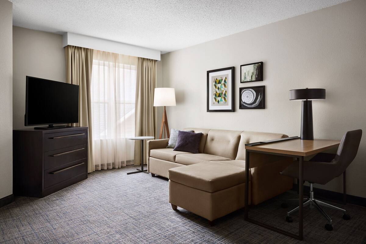 A hotel room with a flat-screen TV, a sofa, a desk, and a chair.