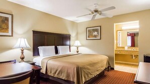 A hotel room with a bed, two bedside lamps, a ceiling fan, a desk with a chair, a small table, a framed picture on the wall, and a bathroom visible through an open door. - Bryant Inn & Suites (Bryant)