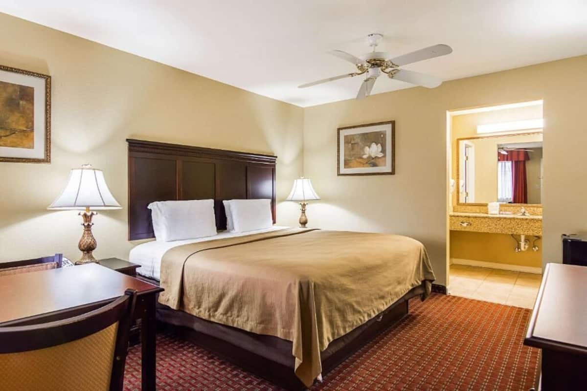 A hotel room with a bed, two bedside lamps, a ceiling fan, a desk with a chair, a small table, a framed picture on the wall, and a bathroom visible through an open door.