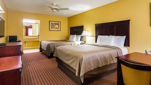 A hotel room with two beds, a desk, a chair, a desk lamp, a ceiling fan, and a painting on the wall. - Bryant Inn & Suites (Bryant)