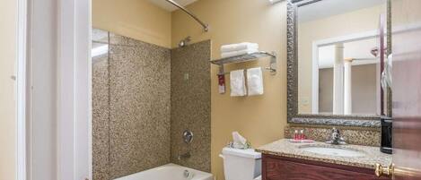 Een badkamer met een bad, toilet en douche. Er is een wastafel met spiegel en een handdoekhouder boven de wasbak.