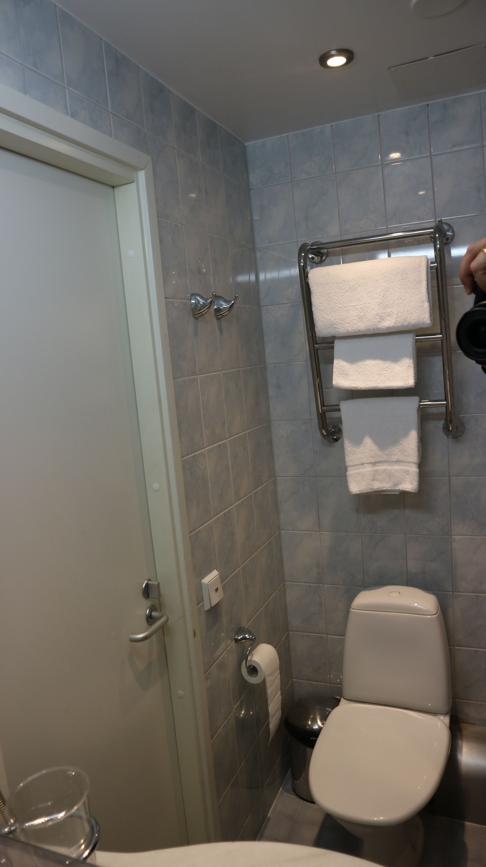A bathroom with a toilet, a towel rack with two towels, a toilet paper holder, and a glass shower enclosure.