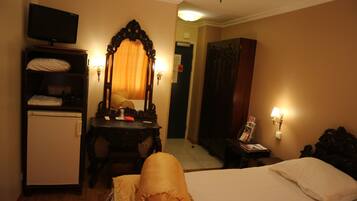 A hotel room with a bed, a desk with a mirror, a television, a wardrobe, and a small table with a lamp.