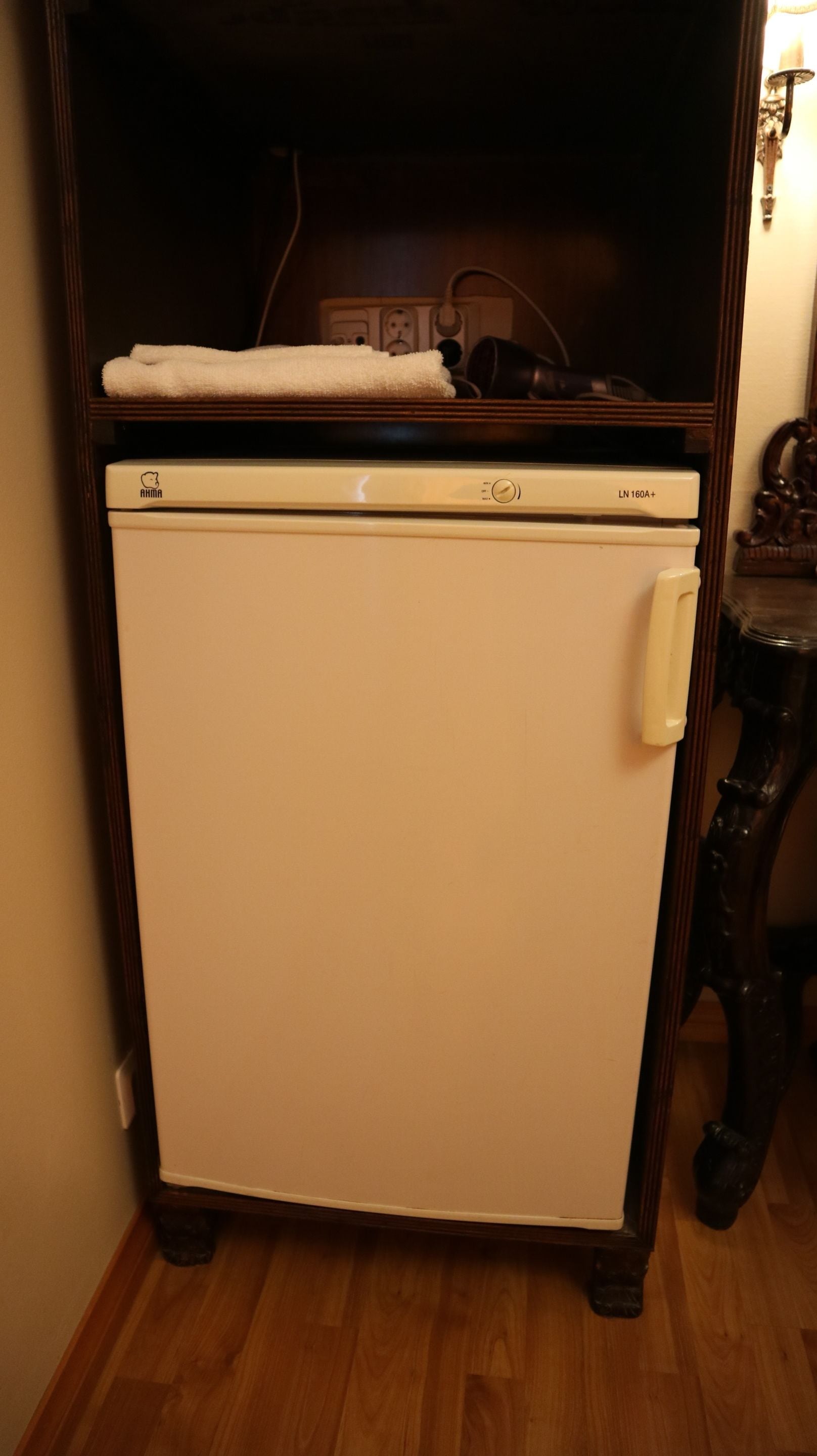 Ein kleiner, weißer Kühlschrank mit Bedienfeld und Griff, platziert in einer Ecke mit Holzboden und einem dunklen Holzschrank darüber.