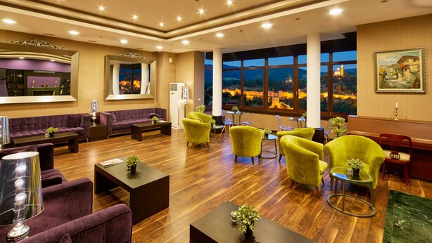 Un hall d’entrée spacieux avec un plancher en bois, un canapé violet, des fauteuils jaunes et une grande fenêtre donnant sur un château illuminé.