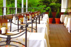 A row of tables with white tablecloths and metal chairs, arranged on a wooden deck with potted plants and a view of greenery.