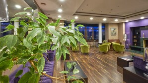 Un hall d’hôtel moderne, agrémenté d’une grande plante d’intérieur, de canapés violets, de fauteuils verts et d’une cheminée.