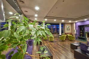 Hall d'hotel moderna con una grande pianta da interno, divani viola, poltrone verdi e un caminetto.