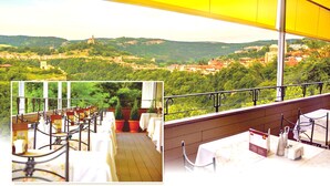 A balcony with tables and chairs, overlooking a scenic view of a town with hills and a church. - Yantra Grand Hotel -sharlopov Hotels (Veliko Tarnovo)