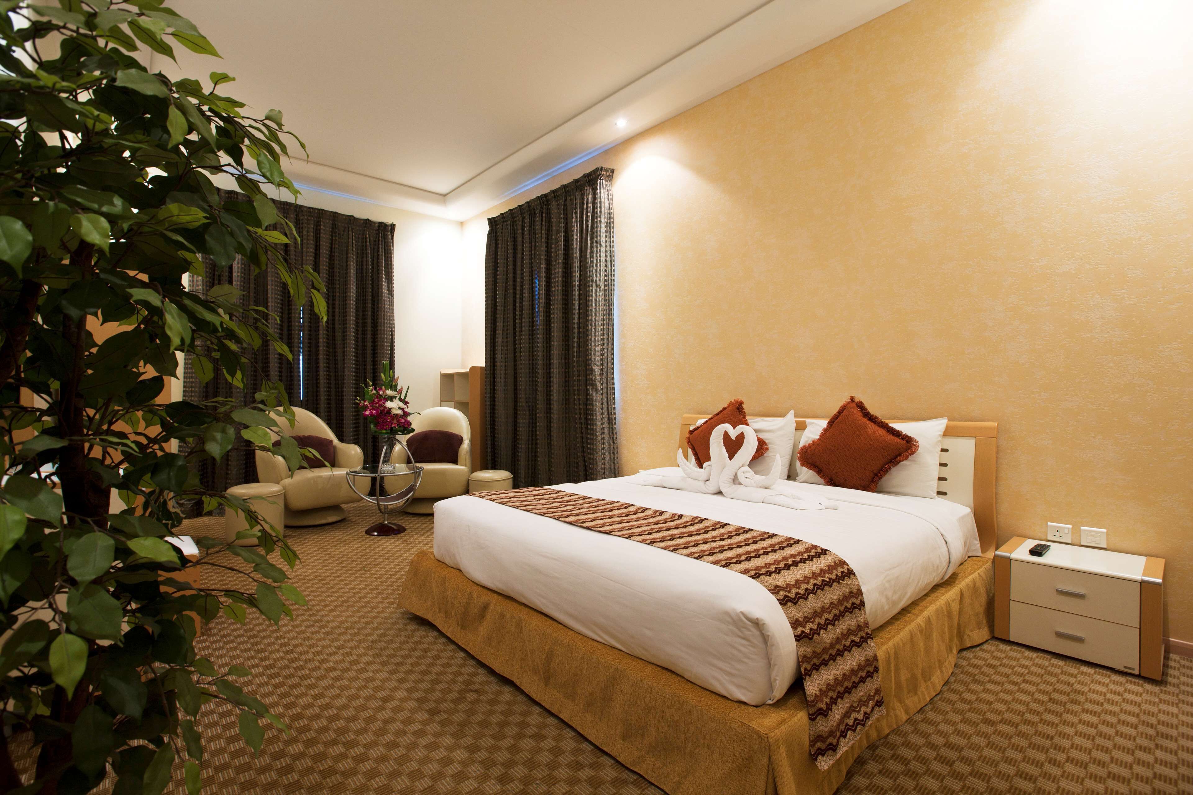 A hotel room with a large bed, a bedside table, a chair, a small table with a vase, and a potted plant.