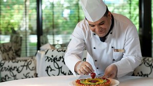 A chef in a white uniform with a name tag, placing a strawberry on a tart on a white plate. - Elite Seef Residence & Hotel (Manama)