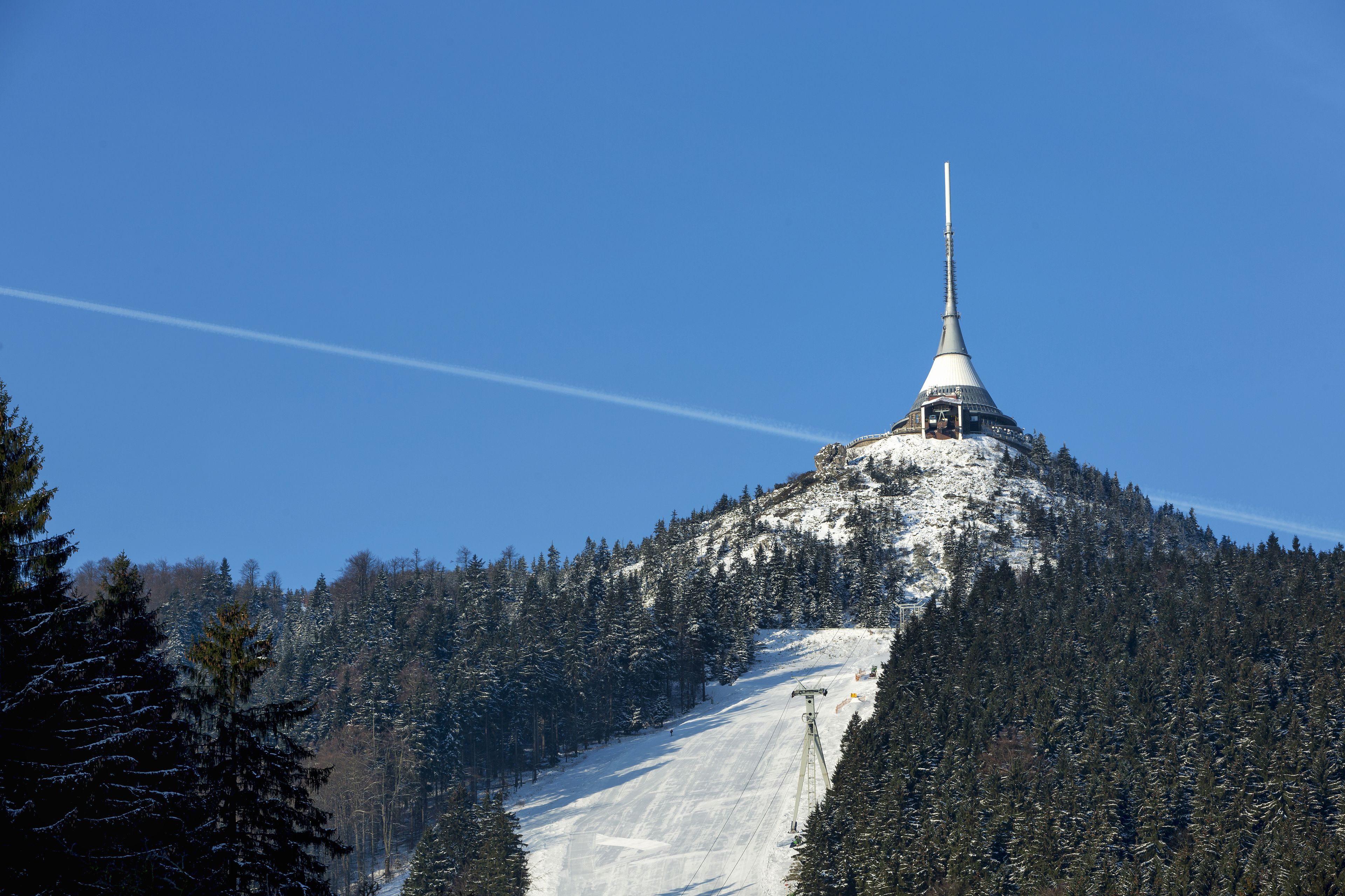 A snow-covered mountain with a tall, pointed structure on top, surrounded by dense forest.