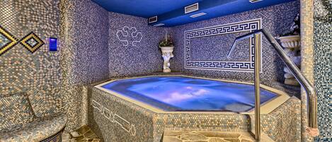 Uma banheira de hidromassagem com paredes de mosaico e piso de pedra, rodeada por um banco de pedra.