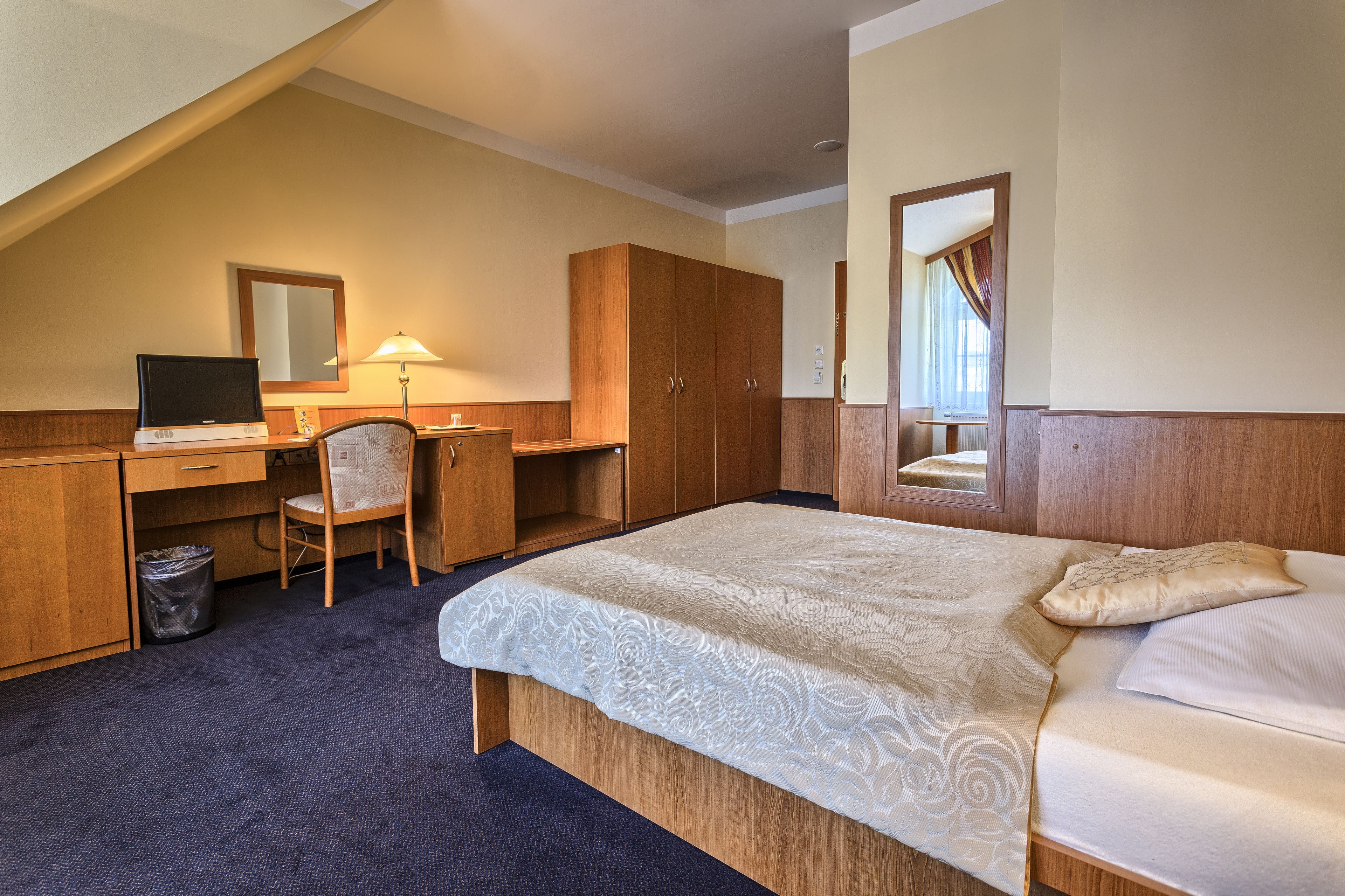 A hotel room with a bed, a desk with a computer, a chair, a wardrobe, and a trash bin.