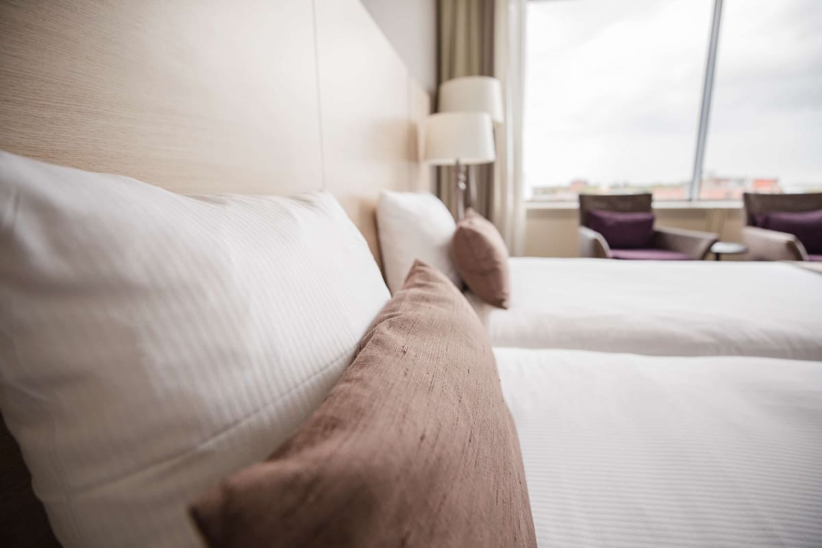 A hotel room with two beds, a lamp, and a view of the city.