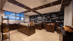 Un área moderna de desayuno tipo bufé con piso de madera, una isla central con alimentos y una pared de pizarra con dibujos y texto relacionado con el desayuno.