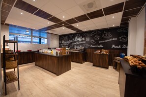 Een moderne ontbijtbuffetruimte met houten vloeren, een centraal eiland met etenswaren, en een prikbordwand met tekeningen en tekst over het ontbijt.