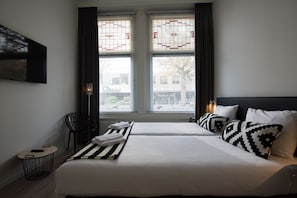 A hotel room with two beds, a flat-screen TV mounted on the wall, a small round table with a chair, and a window with decorative glass panes.