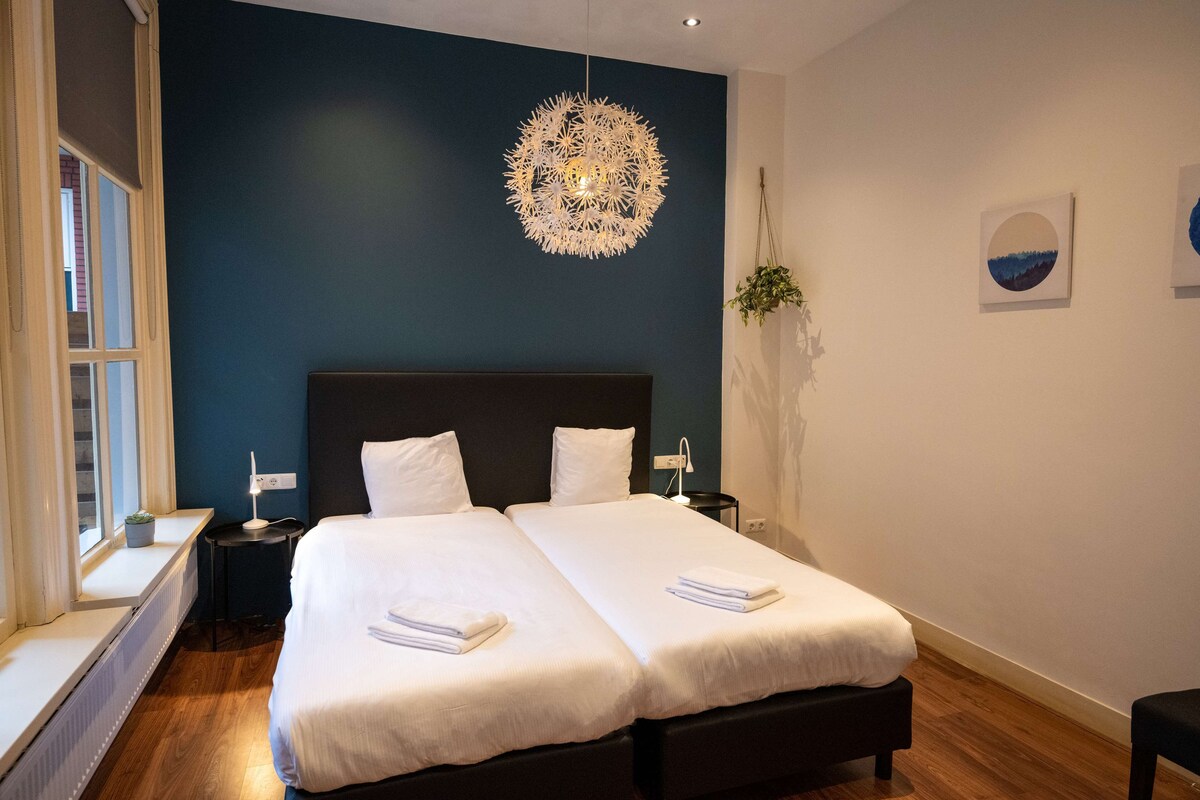 A double bed with white linens, a decorative ceiling light, and a window with a plant on the sill.