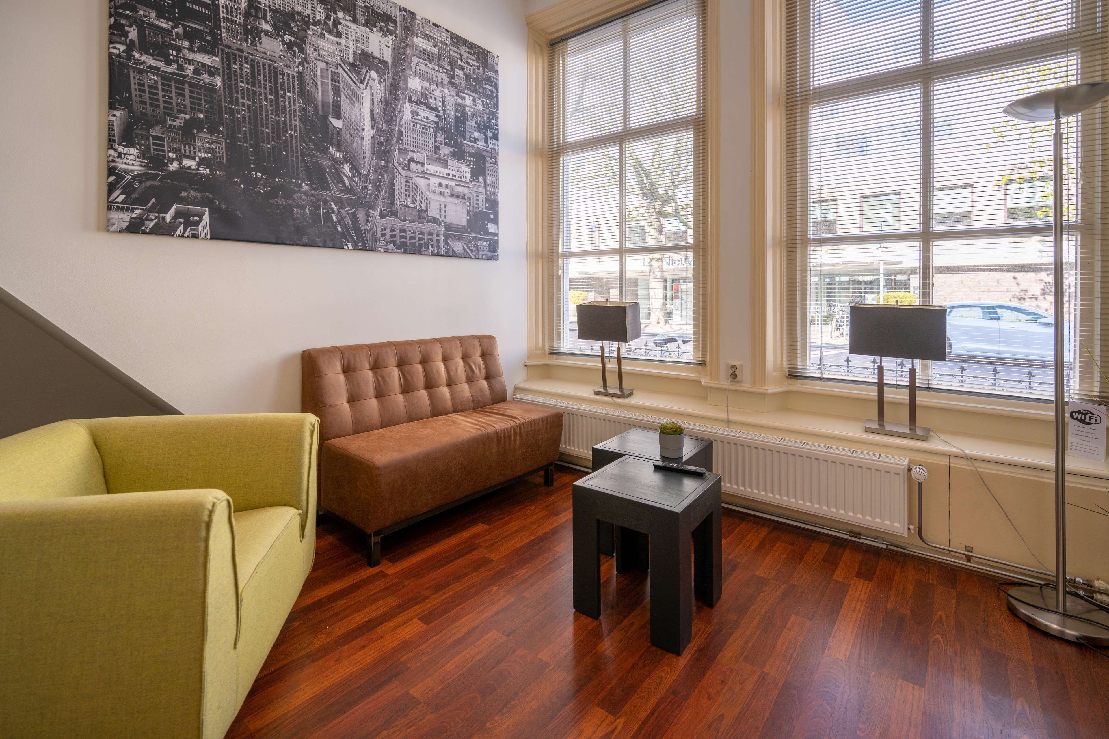Ein modernes Wohnzimmer mit einem braunen Sofa, einem gelben Sessel und einem kleinen Tisch. Es gibt ein großes Fenster mit Jalousien, eine Heizung und ein Stadtbild an der Wand.