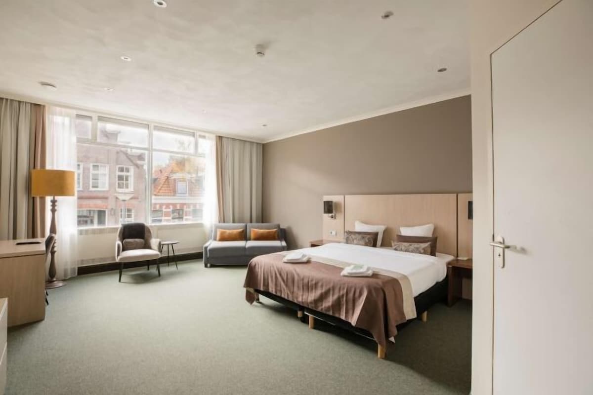 Une chambre d’hôtel équipée d’un lit, d’un bureau, d’une chaise et d’une fenêtre donnant sur des immeubles.