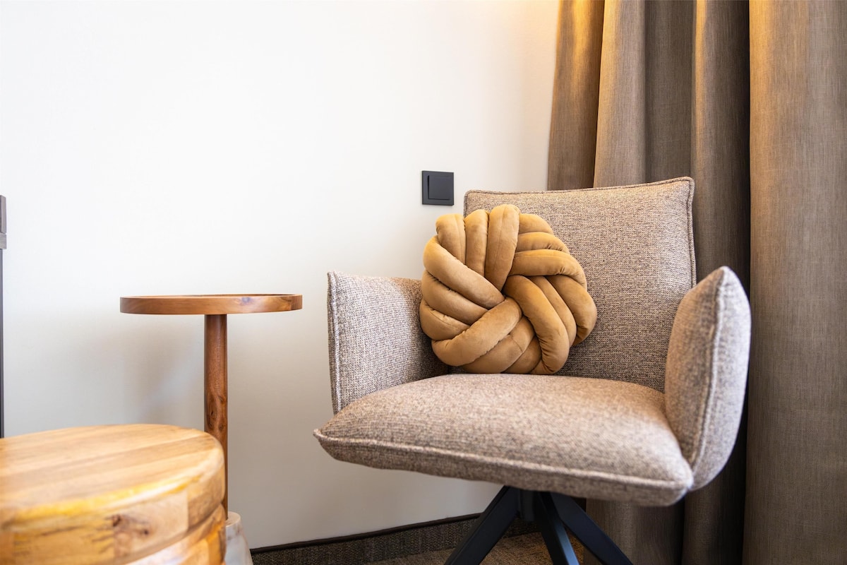 A modern armchair with a unique knot design on the backrest, positioned next to a small wooden table and a wall-mounted light switch.