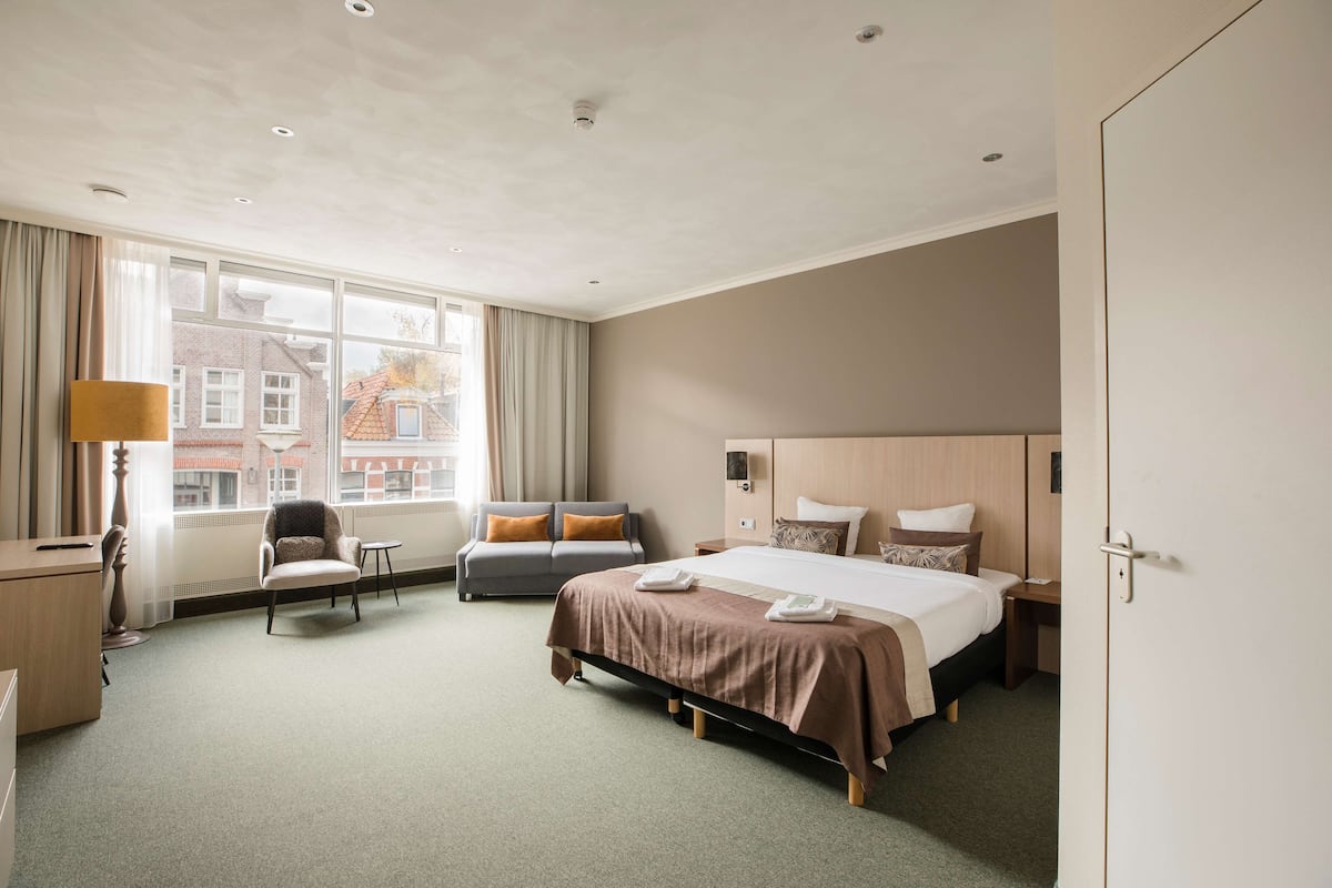 A hotel room with a large bed, a desk, a chair, and a window with a view of buildings.
