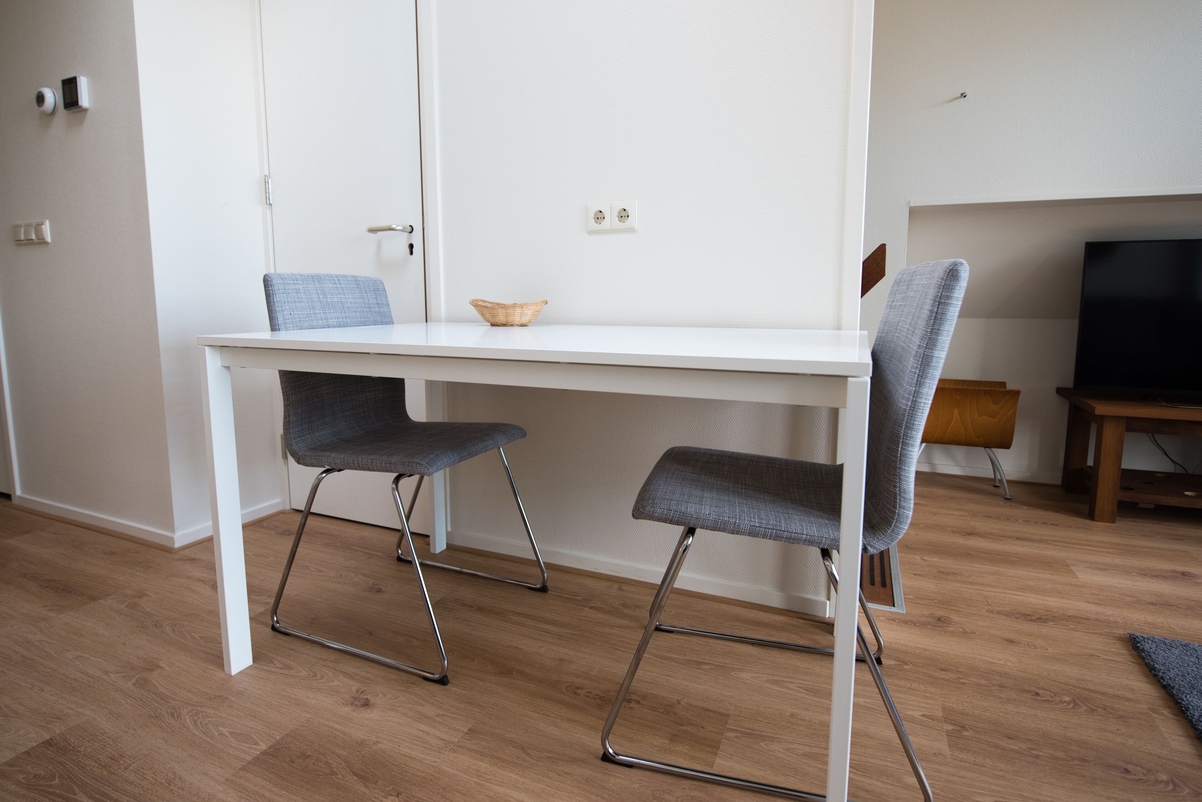 Una sala moderna con una mesa de comedor blanca, dos sillas tapizadas en gris, una pequeña mesita de madera y un televisor sobre un mueble de madera.