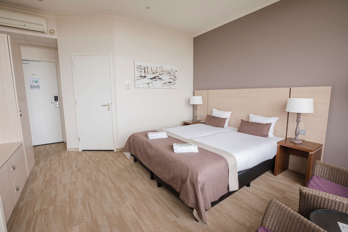 A hotel room with a large bed, two bedside tables with lamps, a chair, and a door leading to another room.
