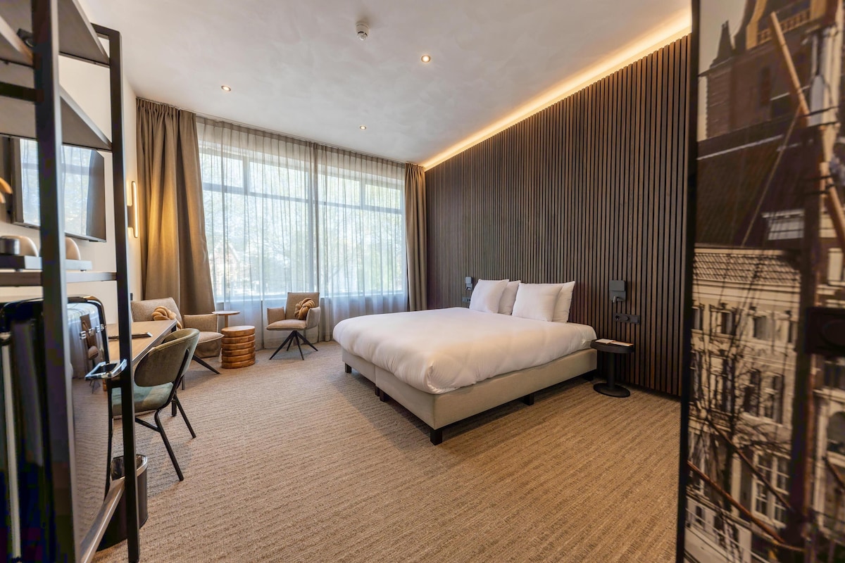 A modern hotel room with a large bed, a desk with a chair, and a view of the city through the window.