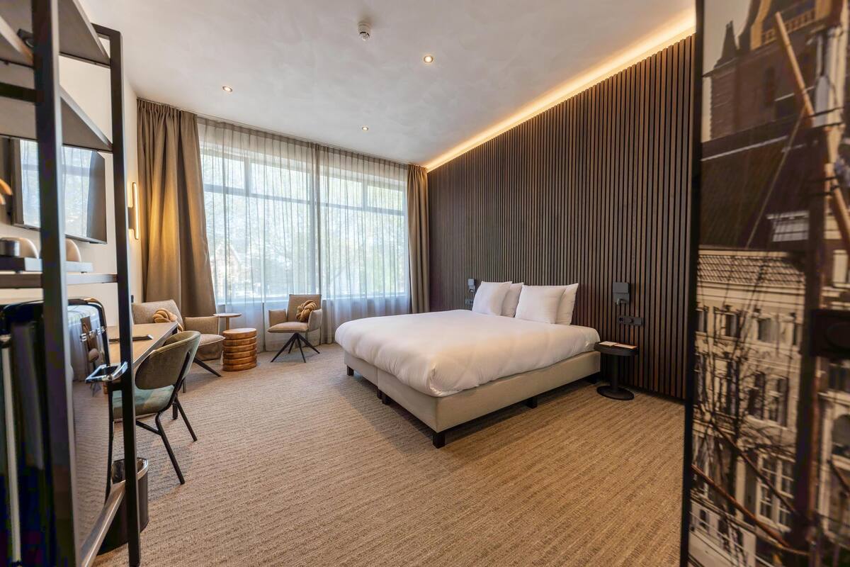 A modern hotel room with a large bed, a desk with a chair, and a view of the city through the window.
