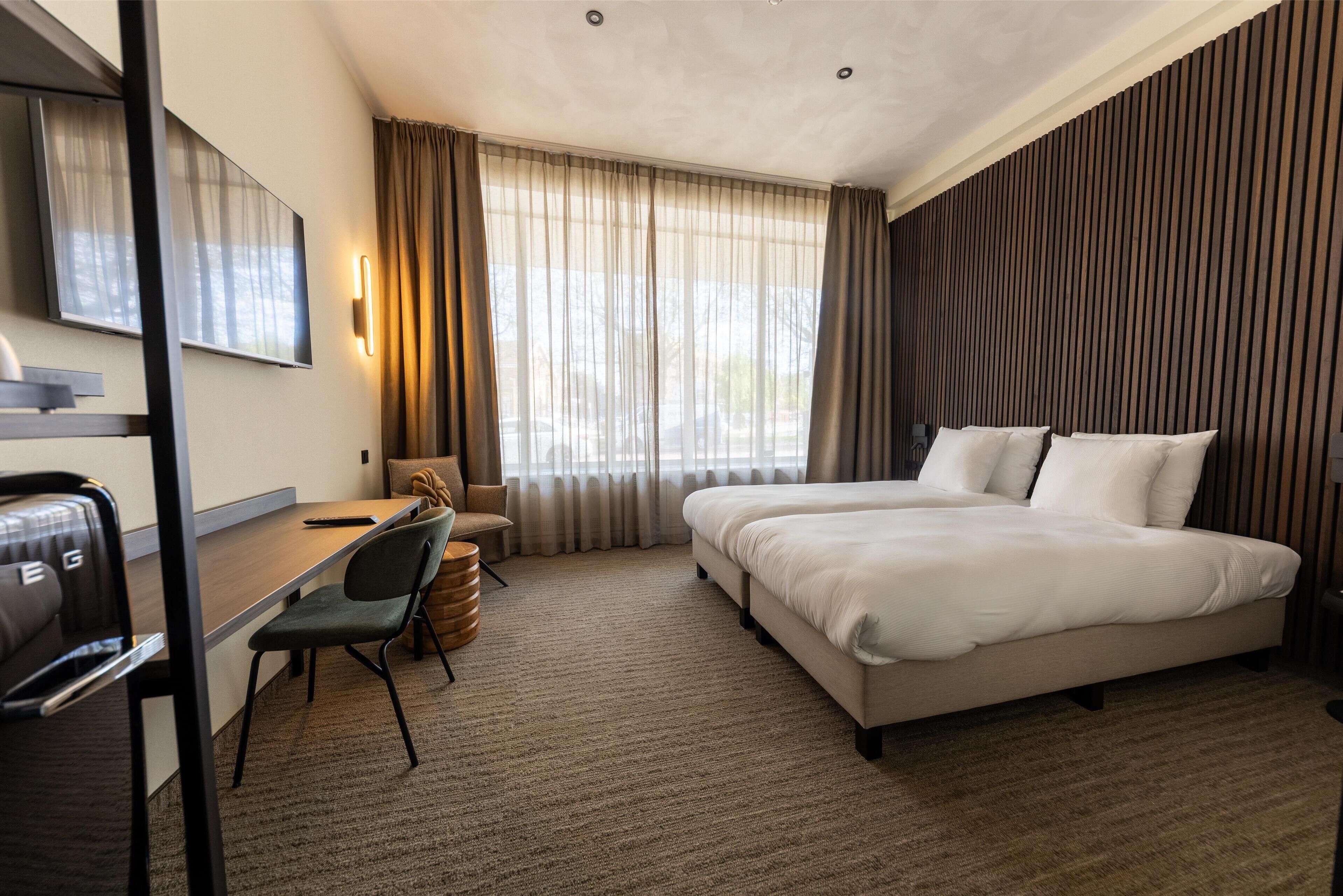 A hotel room with a bed, desk, chair, and a large window with curtains.
