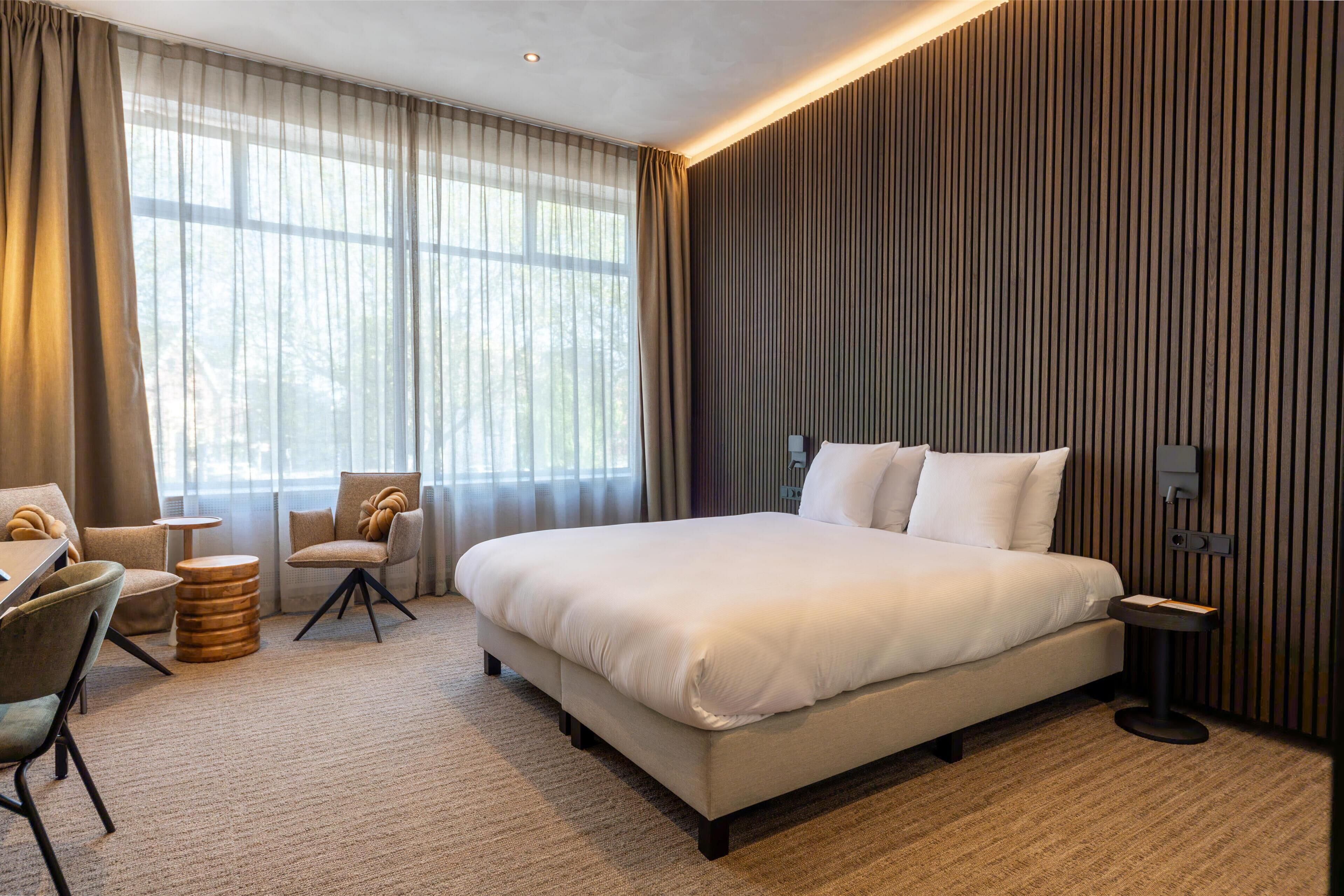 A modern hotel room with a large bed, a seating area with chairs, a small round table, and a large window with curtains.
