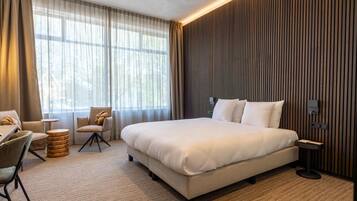A modern hotel room with a large bed, a seating area with chairs, a small round table, and a large window with curtains.
