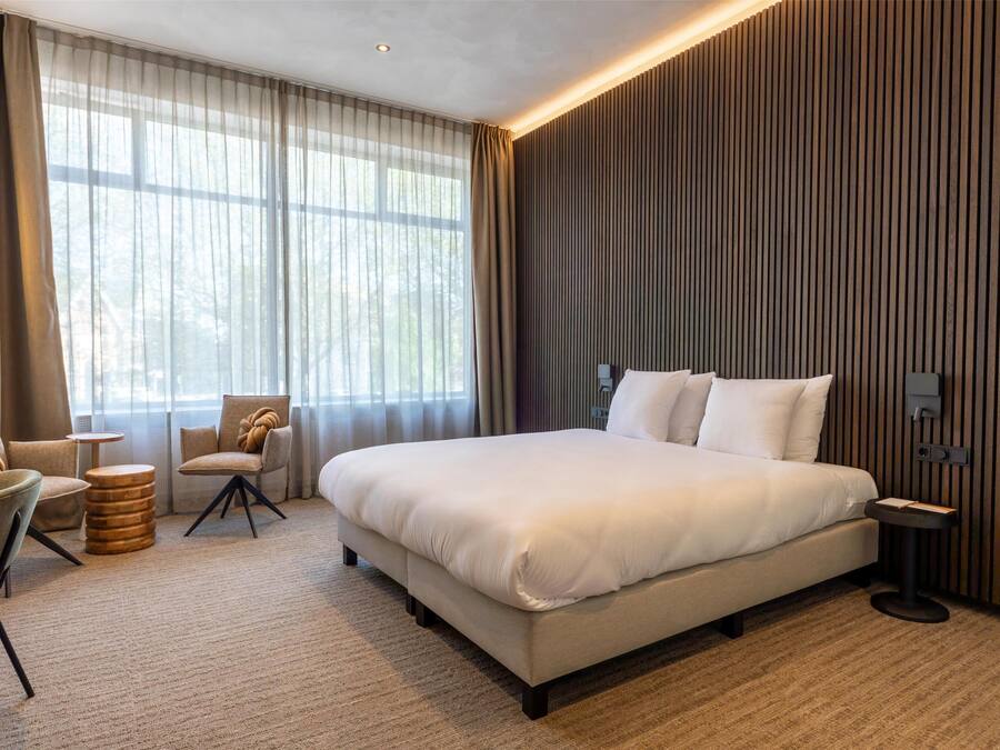 A modern hotel room with a large bed, a seating area with chairs, a small round table, and a large window with curtains.