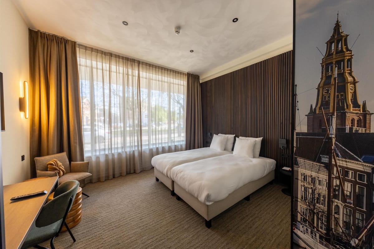 A hotel room with a large bed, a desk with a chair, and a view of a cityscape through the window.