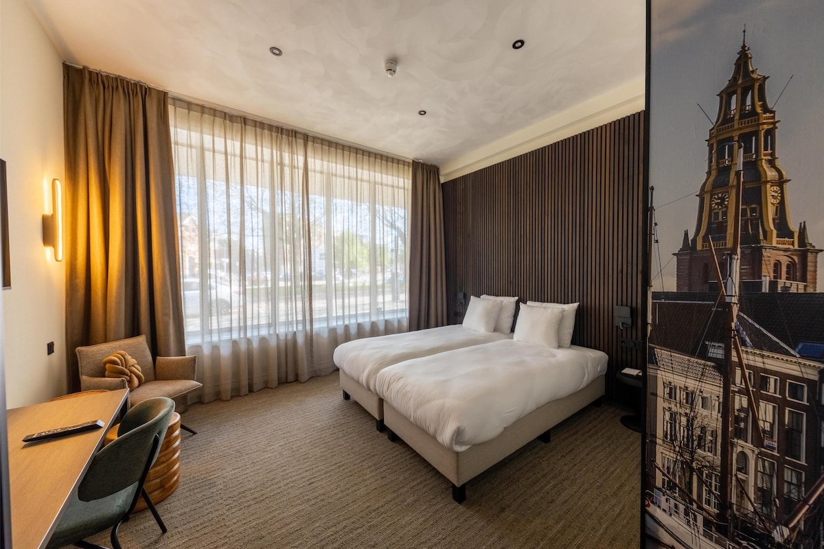 A hotel room with a large bed, a desk with a chair, and a view of a cityscape through the window.